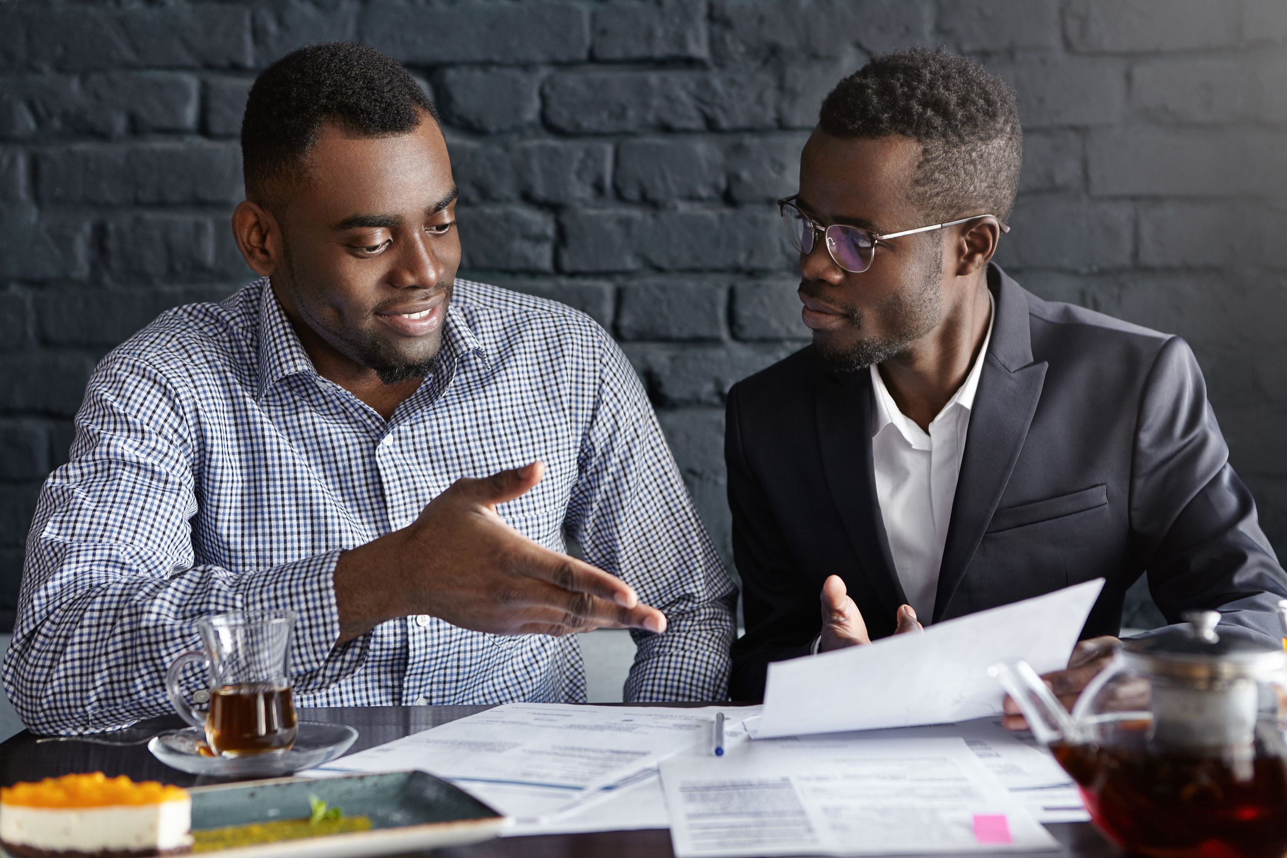 Attractive African businessman in glasses and suit holding papers, showing presentation of his project to cheerful business partner, who smiling happily, supporting his idea with positive gesture